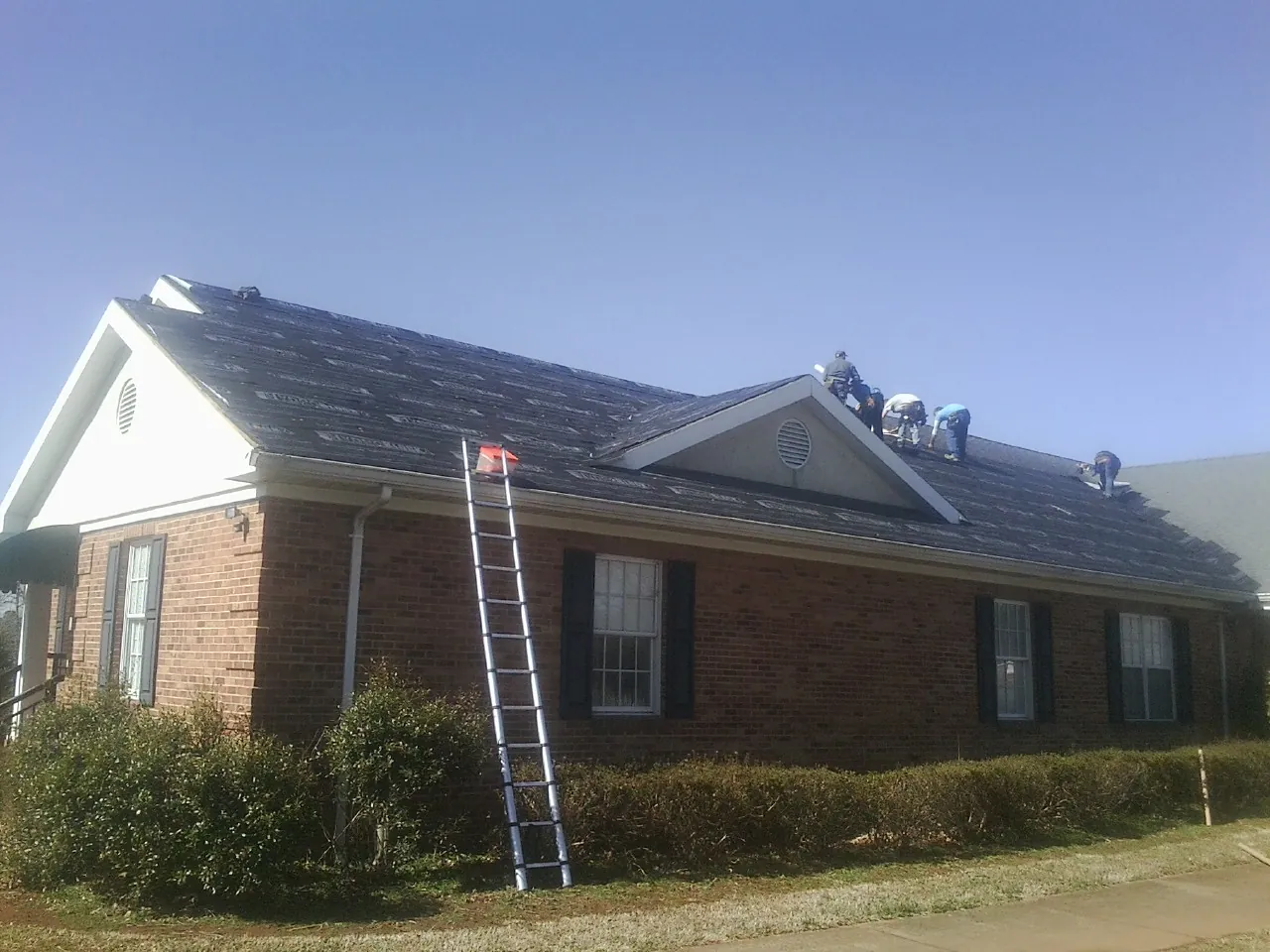 Crew tearing shingles off a brick ranch roof with a ladder set — Metal Roof Repair work by our crew in Tate