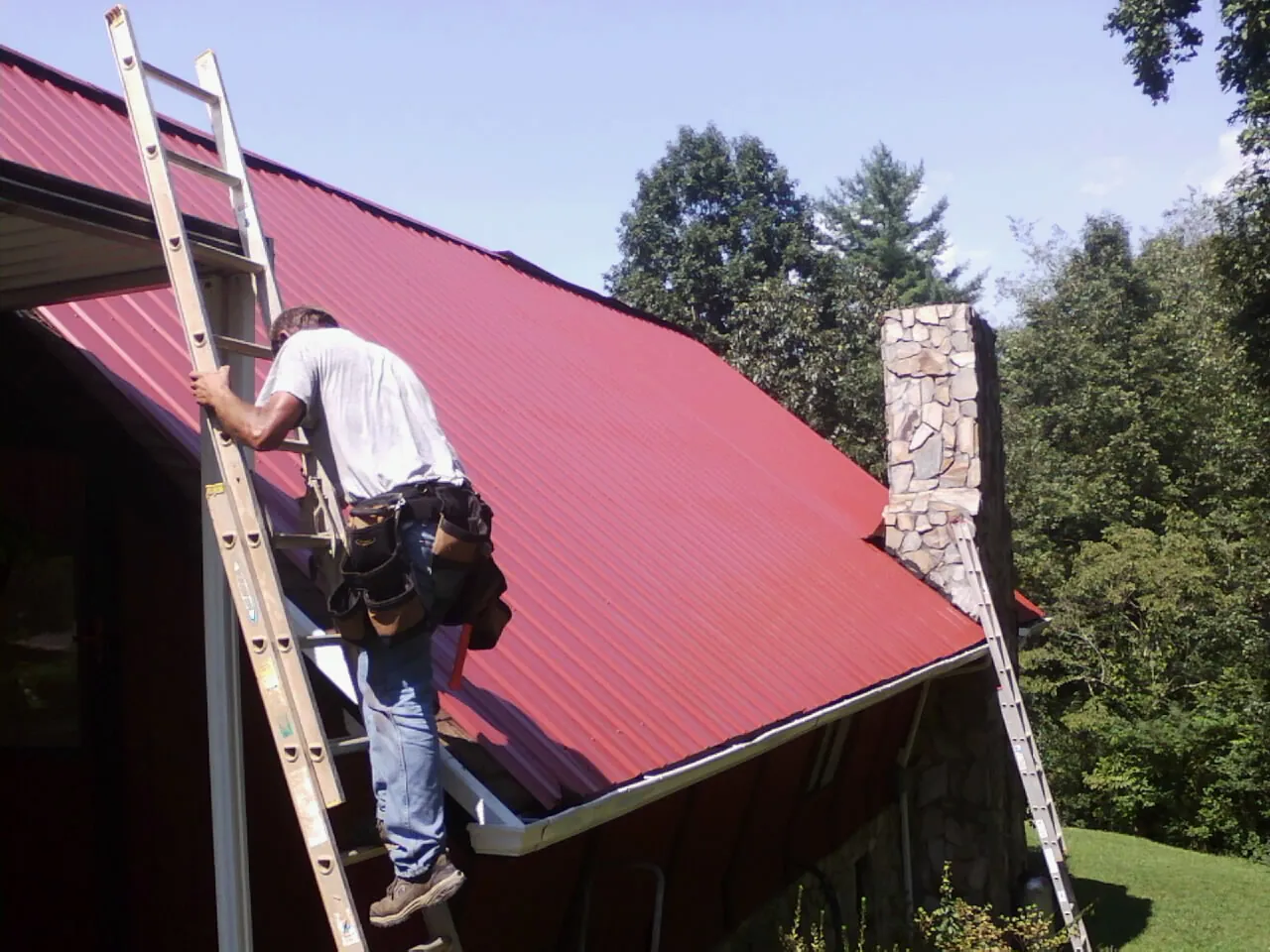 Roofer on a ladder beside a red metal roof and stone chimney — Soffit & Fascia Repair by Roofing Brothers in Tate